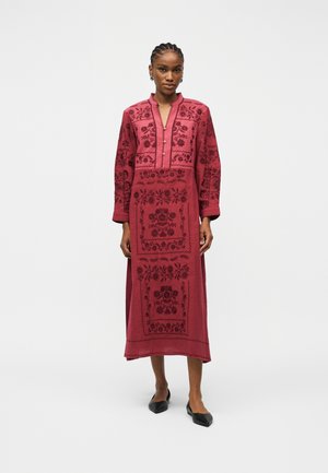 Kobieta stojąca w długiej, czerwonej sukience z ciemnym kwiatowym haftem, długimi rękawami, kołnierzem typu mandarynka i czarnymi, szpiczastymi balerinami.