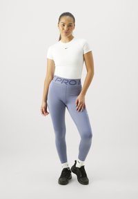Mlada žena ubijena u bijelom pripijenom topu s kratkim rukavima i plavim Nike Pro tajicama, s crnim tenisicama, stoji pred jednostavnom svijetlom pozadinom.