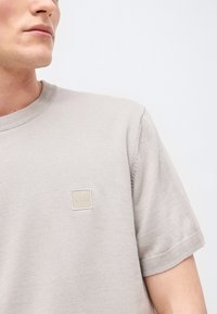 Άνδρας που φοράει ελαφρύ μπεζ πλεκτό πουκάμισο με κοντά μανίκια και τετράγωνο λογότυπο BOSS στο στήθος, κοιτάζει προς τα αριστερά μπροστά σε απλό φόντο.