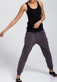 Personne portant un haut noir sans manches et un pantalon gris ample, debout les jambes écartées, le bras gauche tendu sur le côté, sur un fond uni.