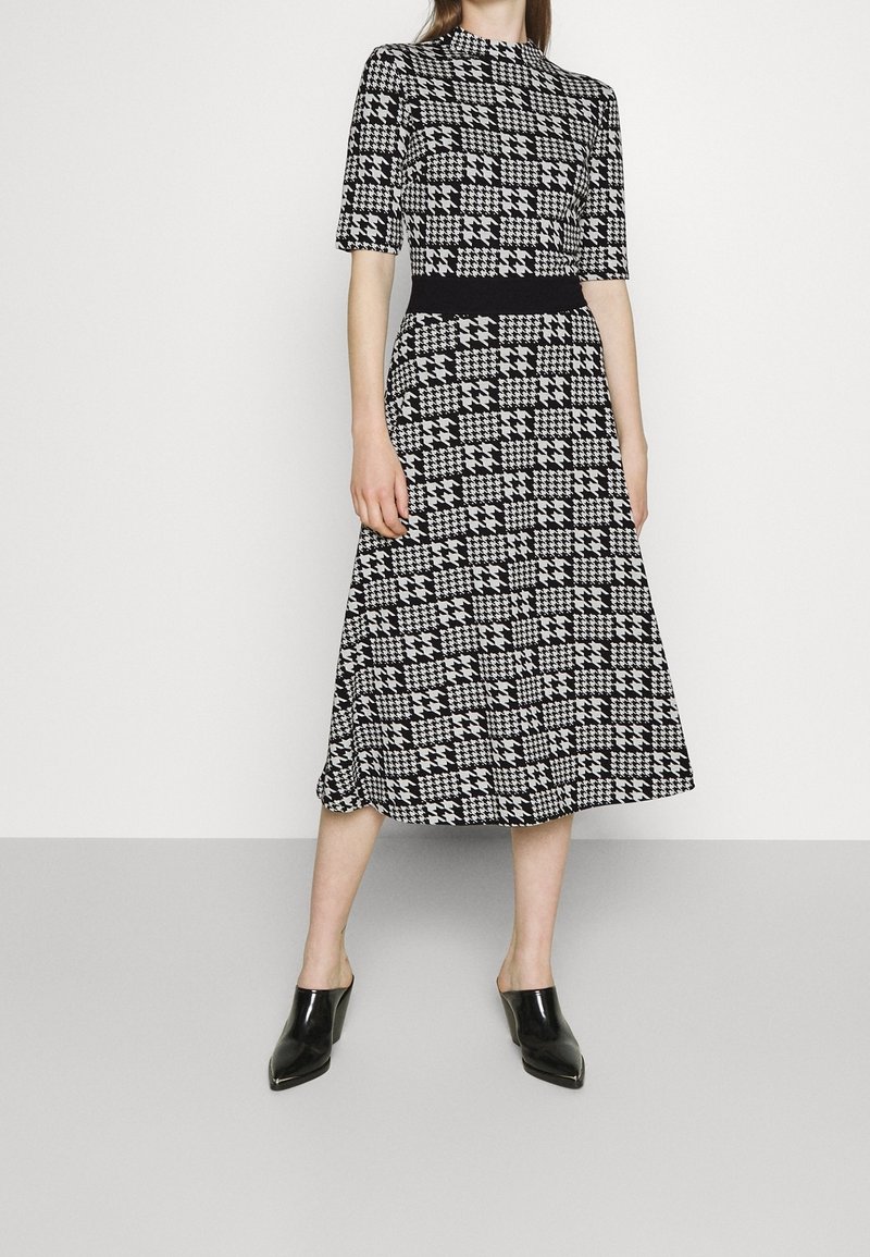 Schwarzes und weißes Kleid im Hahnentritt-Muster mit tailliertem Oberteil, kurzen Ärmeln und einem ausgestellten Midi-Rock. Paar schwarze Mules.