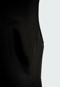 Gros plan d'un vêtement en tissu noir montrant une couture latérale avec un petit détail de poche sur un fond gris clair.