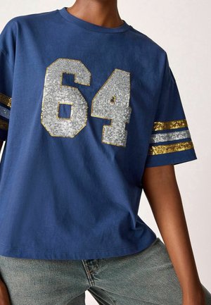 Person trägt ein blaues Kurzarmhemd mit der silbernen Paillettenzahl "64" und gold-silbernen Paillettenstreifen an den Ärmeln, kombiniert mit blauen Jeans.