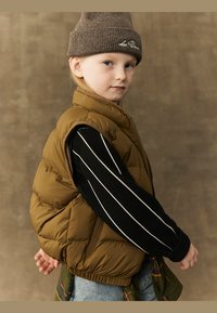 Barna puffasztott mellény, fekete pulóver fehér csíkkal, farmernadrág és barna kötött sapka. Jellemzője a texturált felület és a laza szabás.