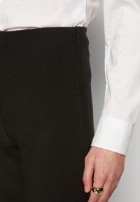 Černé kalhoty s přizpůsobeným střihem, hladkým povrchem a bočním zipem; spárované s bílou košilí a zlatým prstenem na ruce.