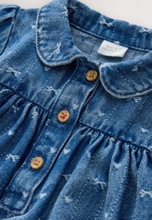 Denimmekko, jossa on sininen kangas, kaulus ja nappikiinnitykset. Mekossa on kerätty tekstuuri ja harvakseltaan valkoisia kuvioita. Napit ovat luonnonpuun sävyiset.