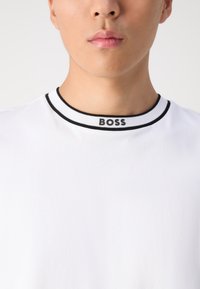 Camiseta blanca con cuello de canalé negro y logo "BOSS". Textura suave, diseño clásico de cuello redondo. Sin patrones ni detalles adicionales.