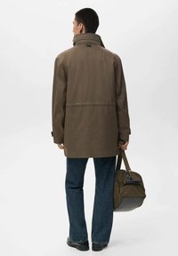 Chaqueta impermeable marrón con un alto cuello, corte holgado y una costura trasera, combinada con jeans de mezclilla oscura y un bolso de hombro verde oliva.