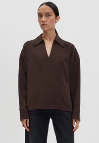 Femme portant un haut ample marron foncé à manches longues avec un large col et un décolleté en V, associé à un pantalon noir, se tenant devant un fond uni.