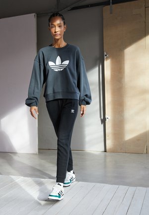 Sudadera negra de gran tamaño con logo blanco, combinada con pantalones jogger negros y zapatillas multicolores, ubicada en una habitación luminosa con luz natural.