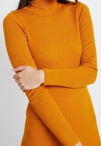 Ribbed golf mundurowy w jasnopomarańczowym kolorze, z długimi rękawami i złotymi guzikami na ramionach jako dodatkowy detal.