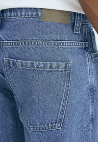 Blå denimjeans med en baklomme, standard søm og en lærlabel på linningen, som viser et klassisk snitt og tekstur.