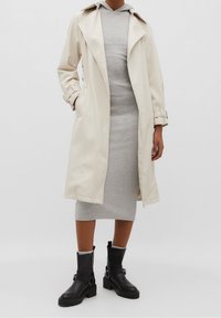 Trench-coat beige avec col, poches latérales et ceinture. Porté sur une robe sweat à capuche grise. Bottines noires avec détails à boucle et semelles épaisses.