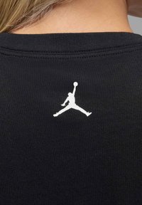 Tela de camiseta negra con cuello acanalado que presenta un logotipo Jumpman blanco en una pose de salto en la parte superior de la espalda.