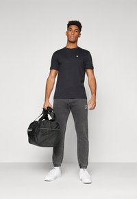 Svart t-skjorte med kortermet og logo, grå joggebukser med elastiske mansjetter, og en svart sportsduffelbag. Hvite joggesko.