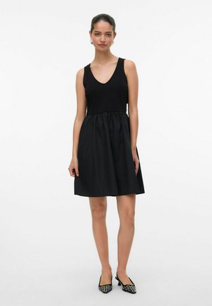Robe noire avec un décolleté en V et sans manches, présentant une taille froncée. Tissu lisse avec une jupe évasée. Portée avec des chaussures noires à motifs.