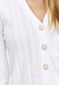 Biała kardiganka z dekoltem w kształcie litery V, teksturowanym wzorem i trzema okrągłymi guzikami z złotymi akcentami z przodu.