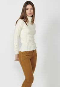 Turtleneck-Pullover in Creme mit geripptem Muster, figurbetontem Design und Knopfdetails an den Ärmeln, kombiniert mit braunen Babycord-Hosen.