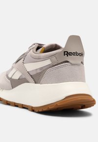Beige och grå sportskor med mocka och tygmaterial, med ett vitt streck, brun sula och svart Reebok-logotyp på hälen.