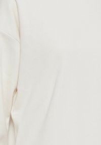 Tessuto di cotone bianco, texture liscia con leggero strech; presenta una vestibilità rilassata e dettagli di cucitura discreti. Primo piano della superficie dell'indumento.