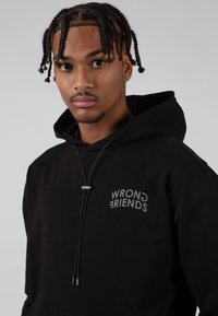 Felpa nera con il logo "WRONG FRIENDS" in grigio. Tessuto morbido con cappuccio regolabile e tasca frontale. Vestibilità casual e rilassata.