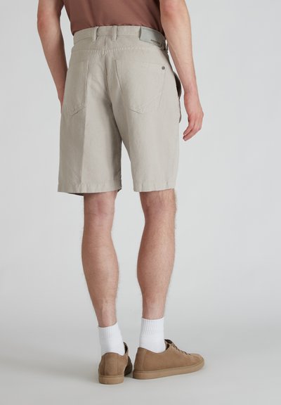Pantalones cortos beige con un diseño liso, dos bolsillos traseros y una etiqueta en la cintura. Llevados con calcetines claros y zapatillas marrones.