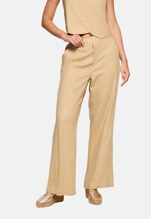 Pantalones anchos beige combinados con un top corto sin mangas con botones y alpargatas de cuña metálicas sobre un fondo blanco.