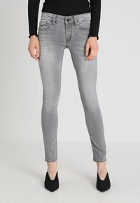 Jeans skinny gris ajustados con una textura desgastada, cinco bolsillos y una cintura con pasadores para cinturón. Llevados con zapatos de tacón alto negros.