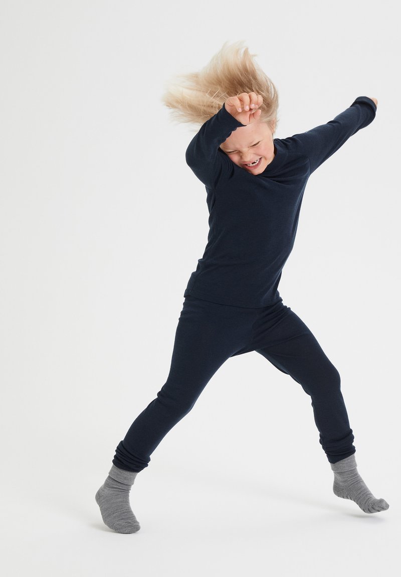 Meriini sininen pitkähihainen paita ja siihen mätsäävät istuvat legginsit, valmistettu pehmeästä kankaasta. Harmaat, ribbikuvioiset nilkkasukat näkyvissä; lapsi dynaamisessa asennossa.