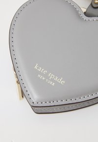 Portafoglio a forma di cuore in pelle grigia con logo dorato "kate spade NEW YORK", cuciture dettagliate e chiusura con zip.