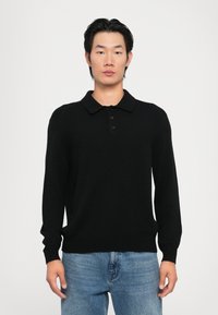 Pulover polo negru cu mânecă lungă, textură cu nervuri, guler cu trei nasturi și design de culoare uniformă. Purtați cu blugi din denim de culoare albastră deschisă.