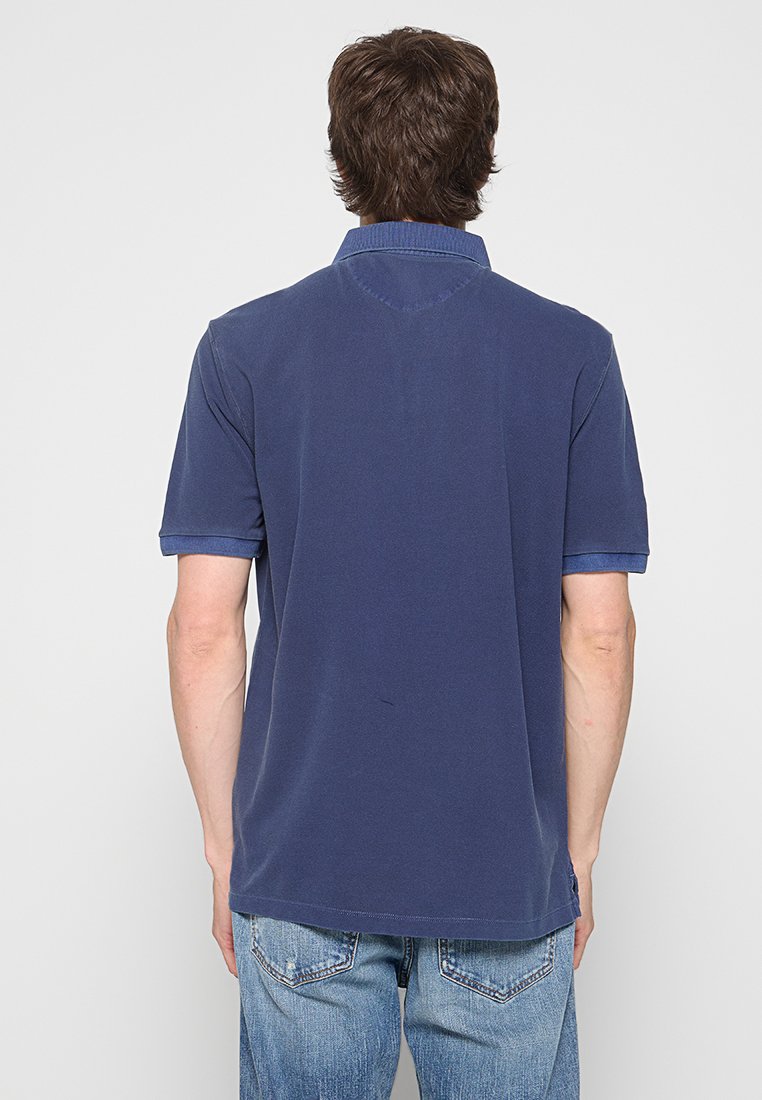 Mężczyzna ubrany w granatową koszulę polo z krótkim rękawem i niebieskie dżinsy, stojący tyłem do kamery na jasnym jednolitym tle.
