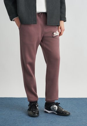 Person, die mauvefarbene Nike-Sweathosen und schwarze weiße Sportschuhe trägt, steht auf einem blau karierten Teppichboden.