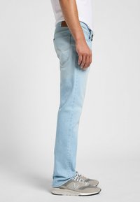 Jeans de mezclilla azul claro con corte recto, bolsillos traseros visibles y un parche de cuero marrón en la cintura. Combinados con zapatillas deportivas grises.