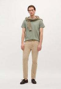 Lys grøn t-shirt, beige bukser og mørke sko. En tan strikket sweater hænger over skuldrene. Simpelt design, glatte teksturer og neutrale farver.