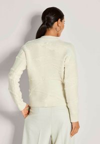 Frau mit dunklen Haaren im Pferdeschwanz, die große Creolenohrringe, einen strukturierten cremefarbenen Pullover und hellfarbene Hosen trägt, steht mit dem Gesicht nach hinten.