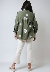 Olijfgroene blouse met witte bloempatronen, lange mouwen, geperste schouders en een ontspannen pasvorm, gecombineerd met crèmekleurige flared broeken en beige sandalen.