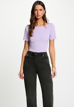 Kvinde står iført en lyse-lilla ribbet kortærmet bluse med knapdetaljer på skuldrene og højtaljede sorte jeans.