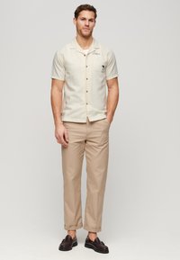 Camicetta corta beige chiaro con chiusura a bottoni, caratterizzata da un design di palme e la scritta "Venice". Indossata con pantaloni beige e scarpe scure.
