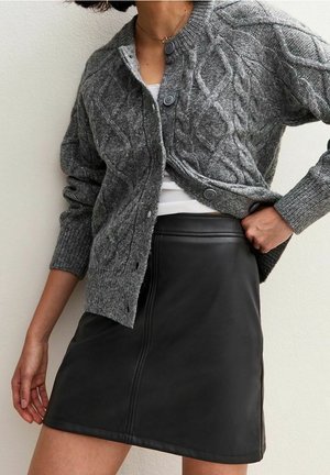 Frau trägt eine graue Zopfmuster-Strickjacke über einem weißen Top und einem schwarzen Leder-Minirock, posiert mit einer Hand an der Hüfte.