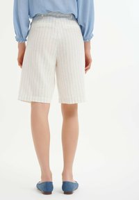 Weiße Leinen-Bermuda-Shorts mit schmalen navy-blauen Streifen, mit geradem Schnitt und umgeschlagenen Säumen, kombiniert mit einem hellblauen Oberteil und blauen Schuhen.