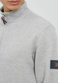 Siva zip-up dukserica s visokim ovratnikom, rebrastom teksturom i kontrastnim plavim krpom na rukavu sa narančastim logotipom.