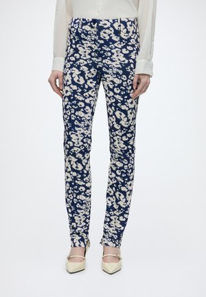 Woman wearing fitted navy blue pants with white abstract floral pattern, cream blouse, and cream pointed-toe heels with ankle straps.