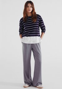 Svetr v tmavě fialové barvě s bílými pruhy, položený přes bílou košili s knoflíky. Šedé kalhoty širokého střihu a světlé boty doplňují celkový vzhled.