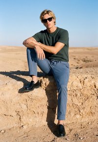Jeune homme portant des lunettes de soleil, un t-shirt vert et un pantalon bleu, assis sur un rebord en pierre dans un paysage désertique sous un ciel clair.