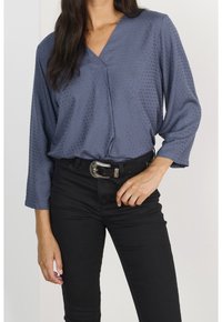 Femme portant un chemisier bleu texturé à col en V avec des manches trois-quarts, rentré dans un pantalon noir taille haute avec une ceinture à boucle argentée.