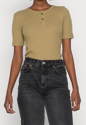 Woman wearing short-sleeve olive green textured henley shirt tucked into high-waisted faded black jeans with hands relaxed at sides.