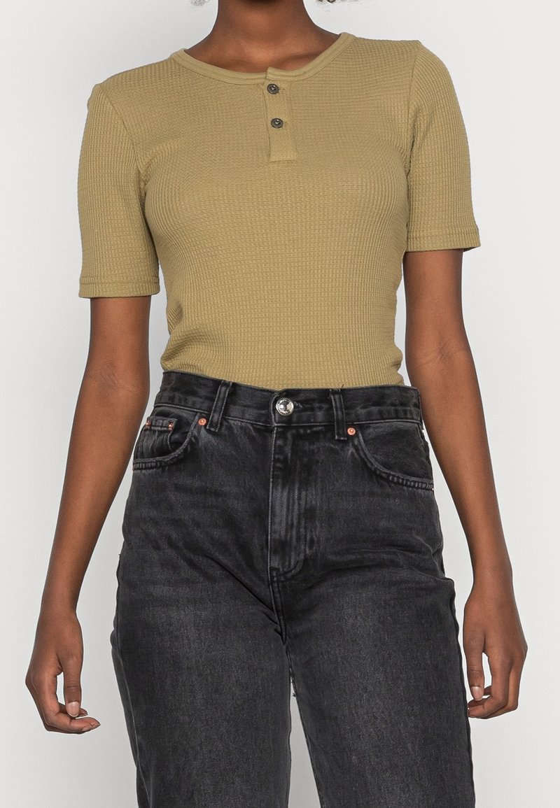 Femme portant un t-shirt henley texturé vert olive à manches courtes, rentré dans un jean noir délavé taille haute, les mains détendues sur les côtés.