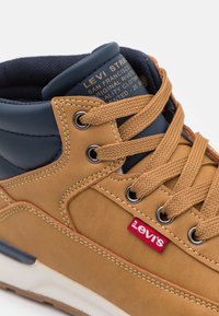 Zapatilla alta de Levi's en tonos marrón y azul marino con cordones marrones, ojales de metal y etiqueta de tela roja con marca en el panel lateral.