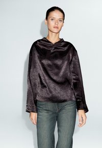 Donna che indossa una blusa di seta satinata lucida di un viola scuro, ampia e con maniche lunghe, insieme a jeans grigi a vita alta, in piedi su uno sfondo bianco semplice.